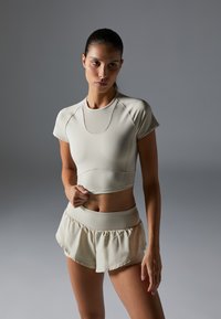 Beige athletic crop top with short sleeves and a round neck, paired with matching high-waisted ruffled shorts; both in a smooth, stretchy fabric.