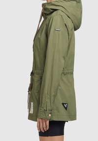 Vista laterale di una persona che indossa una giacca a collo alto verde oliva con cintura regolabile e bottoni, sopra pantaloni neri, in piedi su uno sfondo semplice.
