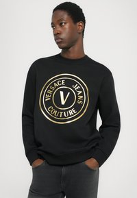 Črna dukserica iz debele tkanine ima okrogel zlat logotip s tekstom "VERSACE JEANS COUTURE." Ribljene manšete in ovratnik.