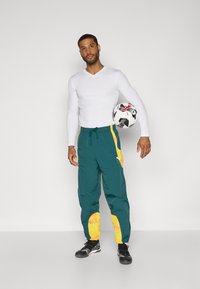Pantaloni sportivi color teal con dettagli gialli alle ginocchia, abbinati a una maglietta attillata a maniche lunghe bianca. Il soggetto tiene in mano un pallone da calcio bianco e nero.