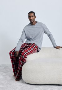 Grå långärmad skjorta och röda rutiga pyjamasbyxor, sittande på en ljusfärgad soffa med rundad struktur mot en blek bakgrund.