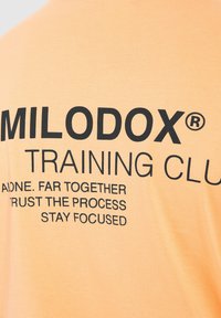 Lys orange bomulds t-shirt med fed sort tekst: "MILODOX TRAINING CLUB" samt motiverende sætninger i forskellige størrelser og justeringer.