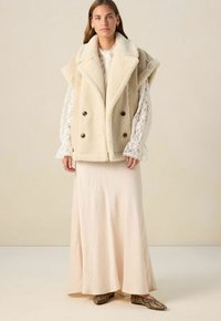 Femme portant un gilet sans manches en fausse fourrure beige, une blouse en dentelle blanche, une longue jupe crème et des ballerines à motif serpent, debout à l'intérieur.