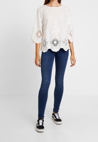 Blusa bianca con design floreale ricamato, orlo ondulato e dettagli in pizzo; abbinata a jeans skinny blu e sneakers nere.