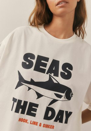 Krémová bavlnená tričko s okrúhlym výstrihom, na ktorom sú výrazné čierne písma a grafika ryby. Text "SEAS THE DAY" je výrazne zobrazený.