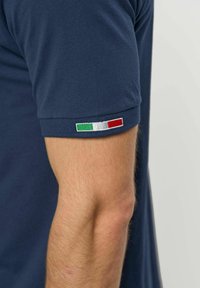 Tengerészkék rövid ujjú ing, kis olasz zászlóval a vállán, egy személy csupasz karját mutatja.