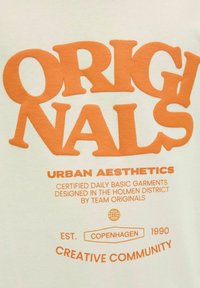 Balts kokvilnas krekls ar izteiksmīgu oranžu tekstu "ORIGINALS" un "URBAN AESTHETICS", ar dizaina detaļām, kas norāda uz izcelsmi Kopenhāgenā, 1990. gadā.