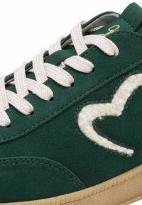 Grønne ruskinds sneakers med cremefarvede snørebånd, prydet med et tekstureret hvidt logo, der ligner uld, og en sål i gummigummi. Glat overflade med syningsdetaljer.