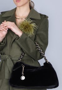 Una donna in un cappotto verde oliva con una spilla a forma di piuma tiene una borsa nera pelosa con una tracolla in oro e un'etichetta rotonda.