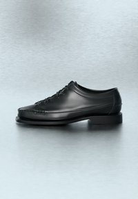 Chaussure habillée en cuir noir à lacets sur le devant avec couture moc toe, présentée sur une surface réfléchissante grise.
