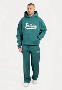 Bluza dresowa i pasujące do niej spodnie dresowe w kolorze morskim, wykonane z miękkiego materiału, z białym napisem z logo na przodzie. Luźny krój, kaptur i kieszenie.