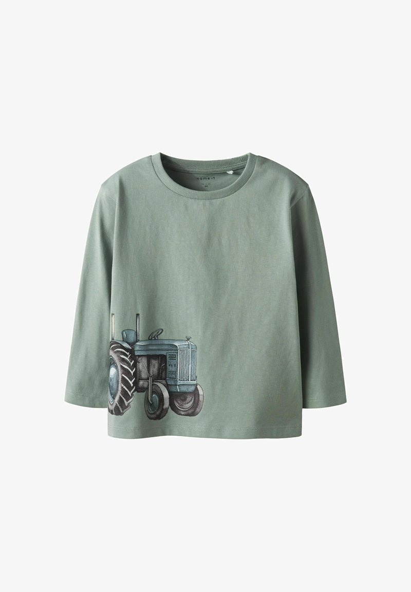 Zaļš garām piedurknēm T-krekls ar apaļu kakla izgriezumu un traktora grafikas printu zilā un pelēkā krāsā apakšpusē. Izgatavots no kokvilnas auduma.