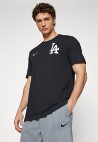 Black cotton t-shirt features a white "LA" graphic and Nike logo. Worn with gray shorts, showcasing a relaxed fit and casual design.