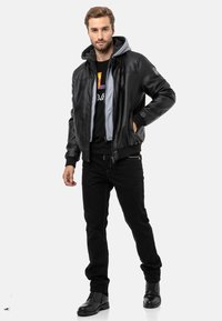 Schwarze Leder-Bomberjacke mit grauem Fleece-gefüttertem Kapuzenpullover, getragen über einem schwarzen Grafik-T-Shirt, kombiniert mit schwarzen Jeans und schwarzen Stiefeln.