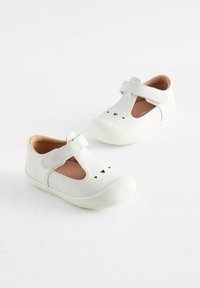 Białe skórzane buty ze wzorem T-strap, gumowymi podeszwami i wycięciami w kształcie serca. Wnętrze jest w kolorze jasnobrązowym, z amortyzowaną wkładką.