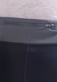 Zbliżenie na ciemnoszare legginsy sportowe z szerokim pasem, który ma poziomą kieszeń na zamek błyskawiczny oraz widoczne szwy na materiale.