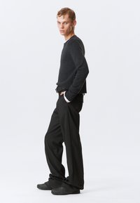 Szary sweter dzianinowy z okrągłym dekoltem, w połączeniu z czarnymi spodniami w paski oraz teksturowanym czarnym obuwiem. Modelka pozuje z boku na tle białym.