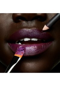 Rossetto viola scuro e lucido su labbra texturizzate, applicato con un pennello. Una matita labbra di una tonalità simile è posizionata nelle vicinanze.