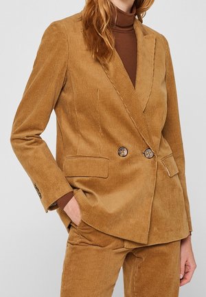 Kvinde iført et kamelfarvet cordjakkesæt med dobbeltradet blazer og matchende bukser, kombineret med en brun rullekravetrøje.
