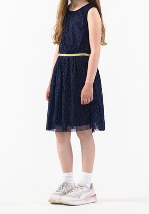 Granatowa sukienka z cekinami, okrągłym dekoltem, bez rękawów, z marszczoną spódnicą i złotym paskiem. W połączeniu z jasnymi trampkami i białymi skarpetkami.