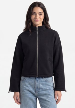 Ung kvinde med langt mørkt hår iført en sort lynlåstrøje og lyseblå jeans, stående mod en ensfarvet hvid baggrund.