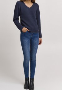 Navyfarbener, strukturierten Strickpullover mit V-Ausschnitt, kombiniert mit schmalen blauen Jeans und schwarzen Ankle-Boots. Verfügt über ein kleines Logo-Detail auf dem Pullover.