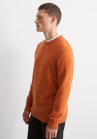 Orange gestrickter Pullover mit Rundhalsausschnitt, gerippten Bündchen und Saum; verarbeitet mit einer strukturierten Oberfläche. Über einem weißen Hemd getragen.