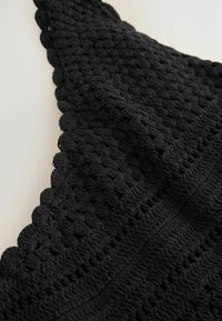 Čierna háčkovaná textília s textúrovaným vzorom, intrikátnym prešitím a vlnitými okrajmi, ktorá preukazuje mäkký a flexibilný materiál.