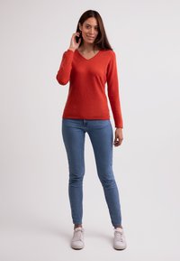 Röd långärmad V-ringad tröja i kombination med blå smalbyxor och vita sneakers. Tyget verkar mjukt med en texturerad yta.