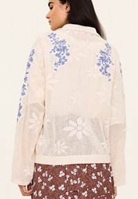 Kevyt kerrosmallinen villatakki, jossa on sinisiä kukka-embroidery olkapäillä, avoin neuletekstuur ja ribbipäälliset, tyylitelty ruskean kukallisen hameen päällä.