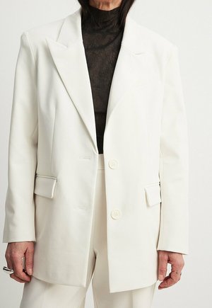 Femme portant un blazer blanc oversize à deux boutons sur un col roulé noir transparent, associé à un pantalon blanc assorti.