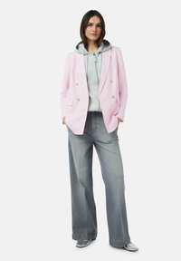 Roze dubbelgeknoopte blazer over een grijze hoodie, gecombineerd met wijde grijze broeken en metallic zilveren instappers. Soepele stof en eigentijds ontwerp.