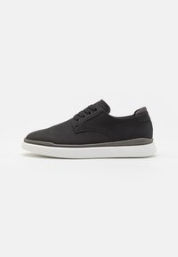 Chaussure noire à lacets avec une tige lisse mate, des accents en mesh texturé et une semelle en caoutchouc blanche. Caractérisée par un bout arrondi et un marquage minimal.