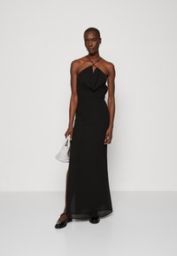 Rochie maxi neagră cu gât halter, cu un corp drapat, siluetă elegantă și fante pe picior. Modelul ține o poșetă mică texturată cu o formă rectangulară.