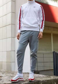 Vit halvdraget jacka med röda ränder, grå byxor med paneldetaljer och vita/röda sneakers, med framträdande Nike-logotyper och branding.