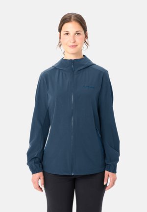 Frau trägt eine marineblaue, mit Reißverschluss versehene Kapuzenjacke mit elastischen Bündchen und schwarze Hose, steht vor einem schlichten hellen Hintergrund.