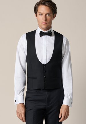 Mężczyzna ubrany w białą elegancką koszulę, czarną muszkę oraz czarną formalną kamizelkę i spodnie, stojący na jednolitym beżowym tle.