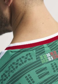 Zelený texturovaný dres s geometrickým vzorem, červeným a bílým lemem a štítkem "SOMOS MEXICO" poblíž výstřihu.