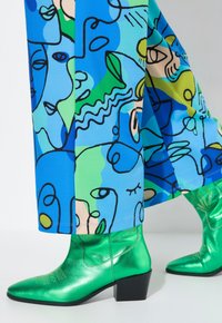 Groene metallic enkellaarzen met een blokhak, gecombineerd met blauwe broek met abstracte gezichten in verschillende tinten blauw en groen.