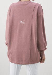 Př oversized růžový fleece svetr s kulatým výstřihem, spadlými rameny a bílým logem Nike AIR na zádech. Hribovaný lem na rukávech.