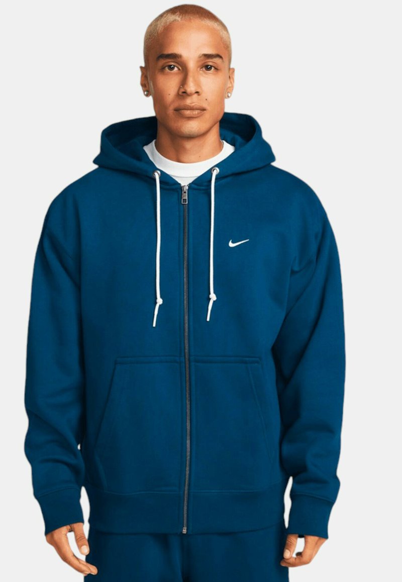 Marinblå zip-up hoodie tillverkad av mjukt material. Har en vit Nike-logotyp, dragsko, kängurufickor och ribbade muddar.
