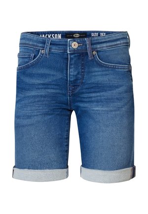 Młoda osoba ubrana w koralowy t-shirt z krótkim rękawem, podwinięte jeansowe szorty, białe skarpetki i białe trampki stoi na białym tle.
