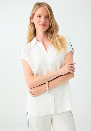 Blondynka o jasnej karnacji, ubrana w białą, koszulę z krótkim rękawem zapinaną na guziki oraz jasne spodnie, stojąca z założonymi ramionami na białym tle.