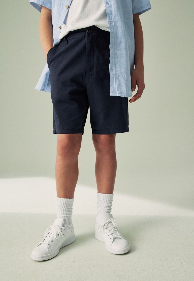 Personne portant un short bleu marine, des baskets blanches avec des chaussettes blanches, un t-shirt blanc et une chemise à manches courtes bleu clair déboutonnée.