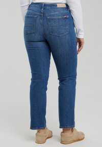 Osoba nosząca niebieskie proste dżinsy z tylnymi kieszeniami, białą koszulę z długim rękawem i beżowe trampki, stojąca na jednolitym tle.