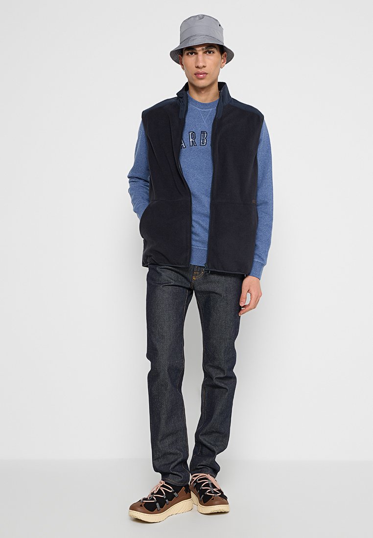 Gilet en polaire bleu marine avec zip à l'avant, associé à un sweat-shirt bleu, un jean en denim sombre et des sneakers marron avec des accents noirs et roses.