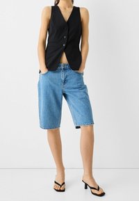 Svart randig väst med fem knappar, parat med ljusblå denimshorts med fransad fåll. Modell som bär svarta sandaletter med remmar.