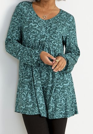 Femme portant une tunique longue à manches longues florale bleu sarcelle avec une taille froncée, associée à un pantalon noir et des bijoux en or aux mains et au cou.