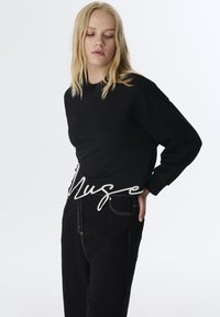 Sudadera negra con escote redondeado y mangas largas. Guía decorativa en blanco con la palabra "Muse" a lo largo de la cintura. Combinada con pantalones negros texturizados.