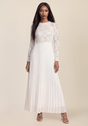 Woman with long straight hair wearing a white lace long-sleeve top and pleated ankle-length skirt standing on beige background.